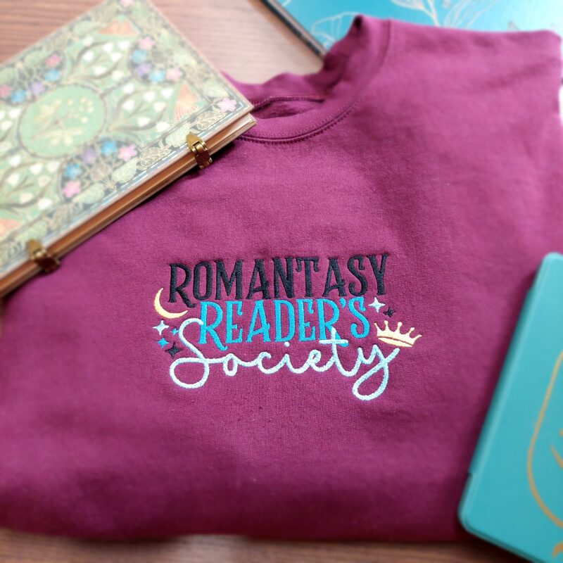 Cozy sweatshirts featuring the phrase "Romantasy Readers Society" prominently displayed on the chest. Perfect for book lovers!