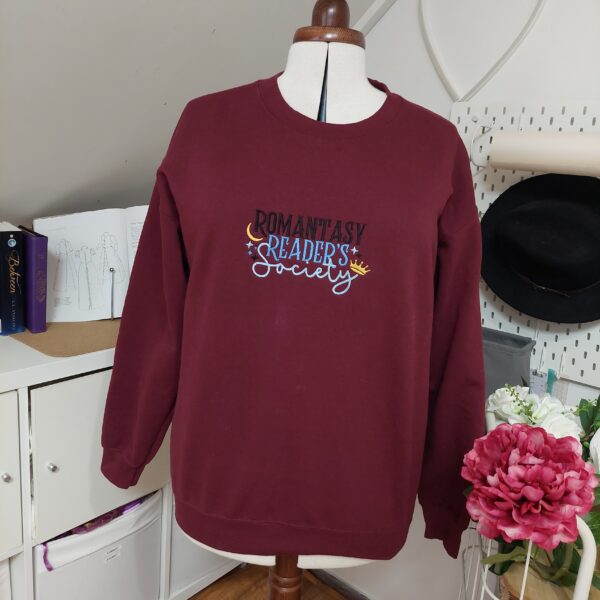 Cozy sweatshirts featuring the phrase "Romantasy Readers Society" prominently displayed on the chest. Perfect for book lovers!